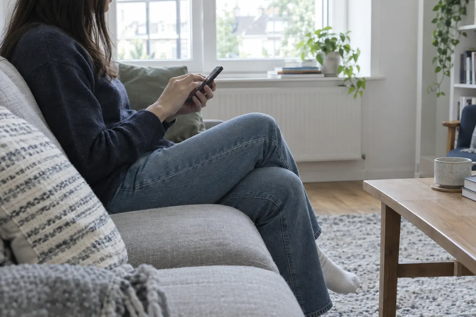 Pessoa sentada num sofá a escrever no telemóvel, numa sala com luz de janela